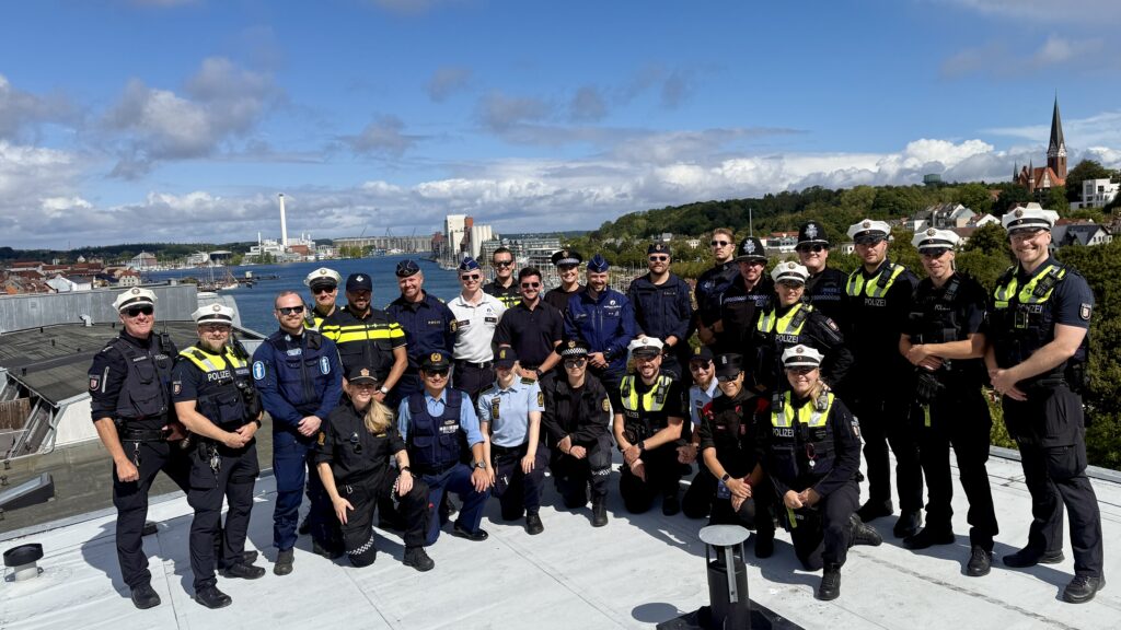 Eine große Gruppe mit unterschiedlichen Uniformen steht auf einem hohen Dach, Im Hintergrund die Flensburger Förde