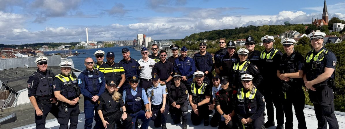 Eine große Gruppe mit unterschiedlichen Uniformen steht auf einem hohen Dach, Im Hintergrund die Flensburger Förde